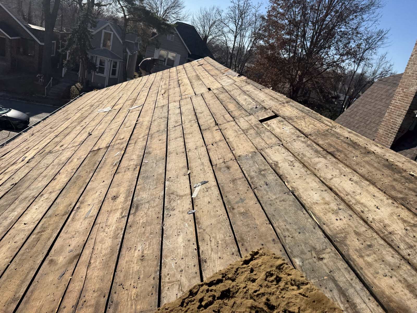 Bare plank decking on a Keys Roofing tear off, photographed up the slope towards the ridge. The boards are weathered and separated by gaps, and the whole surface is covered in the shanks and heads of old roofing nails left behind by the shingles that were stripped from it, with scraps of felt and a pile of granules at the lower right.