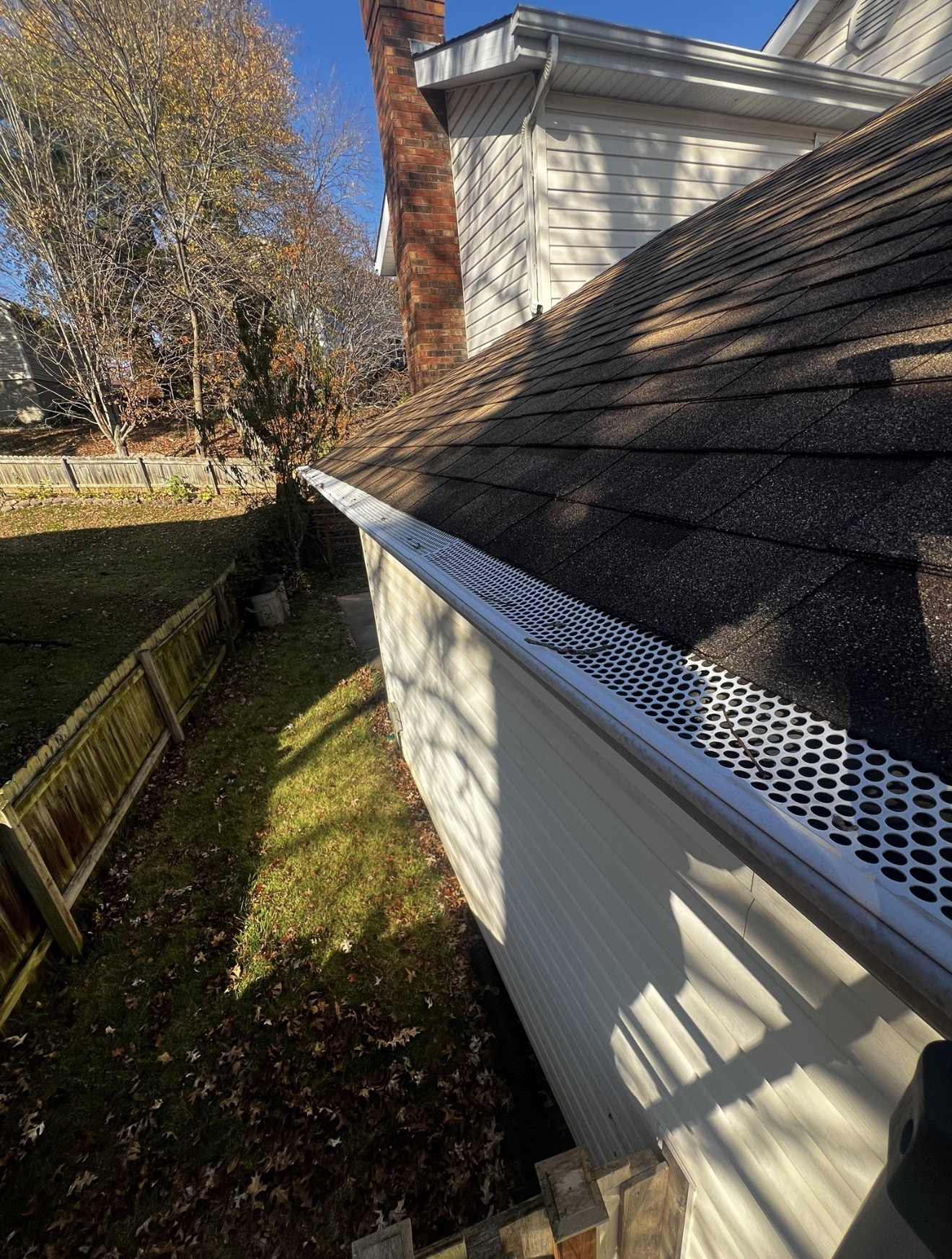 Looking along the eave of a weathered dark shingle roof on a Keys Roofing job, with a white perforated metal gutter guard running the length of the gutter and two bare twigs and a few small dark scraps of debris lying on top of the perforations, a red brick chimney and white vinyl siding above, and a back yard below with a weathered timber fence and fallen autumn leaves on the grass under a clear blue sky.
