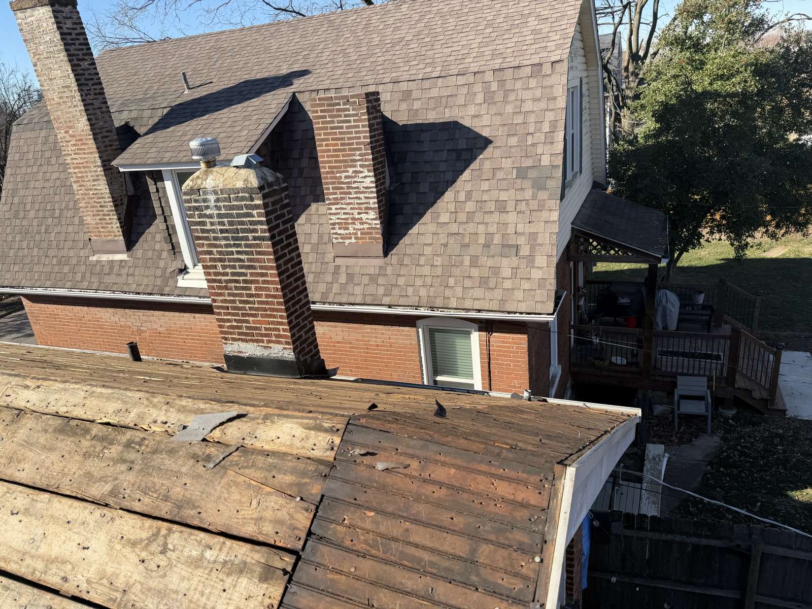 Photographed from a lower roof plane on a Keys Roofing job that has been stripped to its bare plank decking, looking across at the main house: a weathered brown shingle roof, two red brick chimneys with spalled faces and patched mortar, a dormer with a metal turbine vent, a raised timber deck with furniture at the right, and bare and green trees under a clear blue sky.