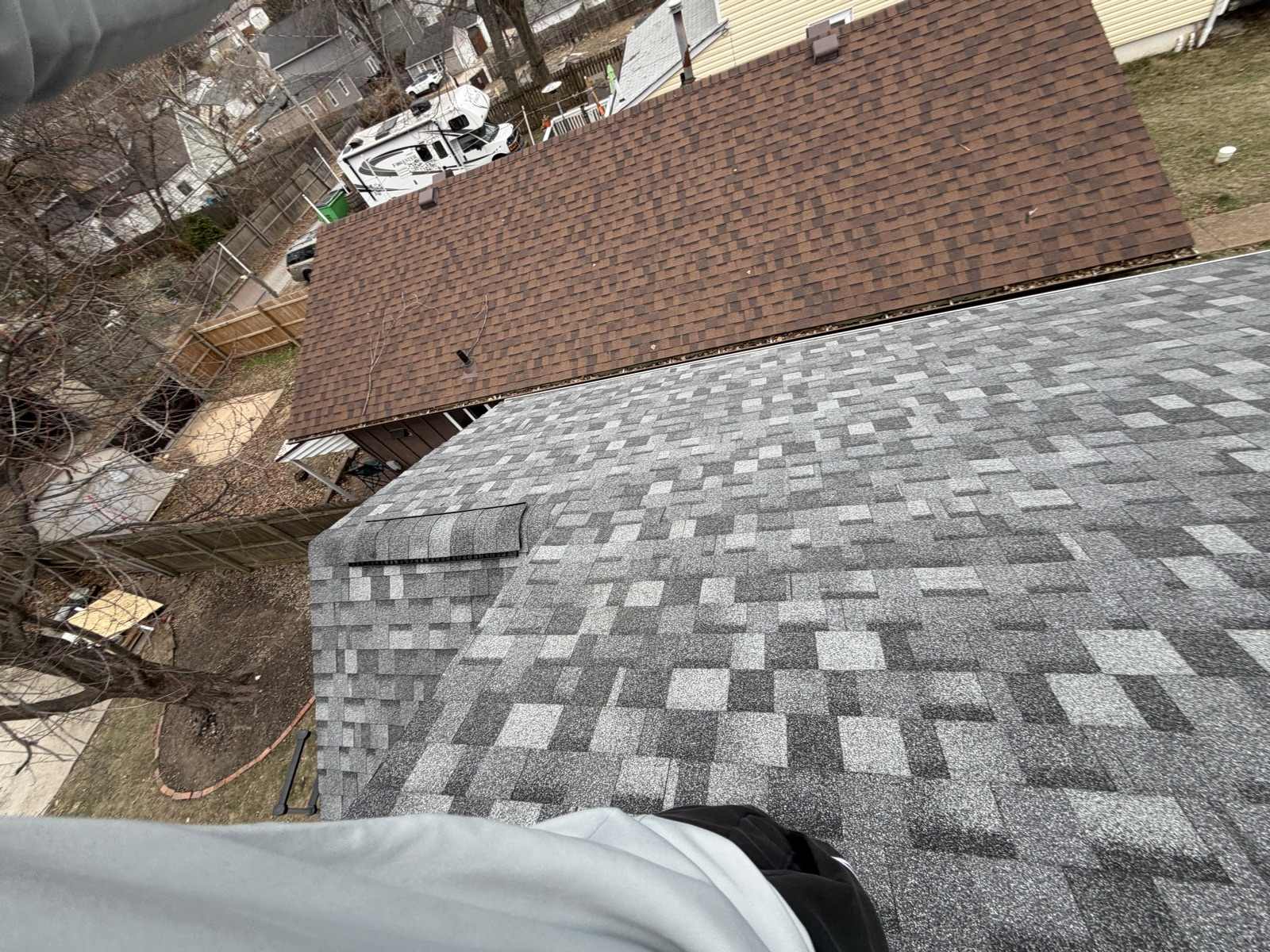 Looking down from the ridge of a completed Keys Roofing job: a new grey and charcoal architectural shingle roof with a ridge vent fills the right and lower part of the frame, and the adjoining older section at the upper left still carries its weathered brown shingles with debris visible in its gutter. Below is a winter back yard with timber fences, a bare tree, a white recreational vehicle in the alley and neighbouring houses beyond.