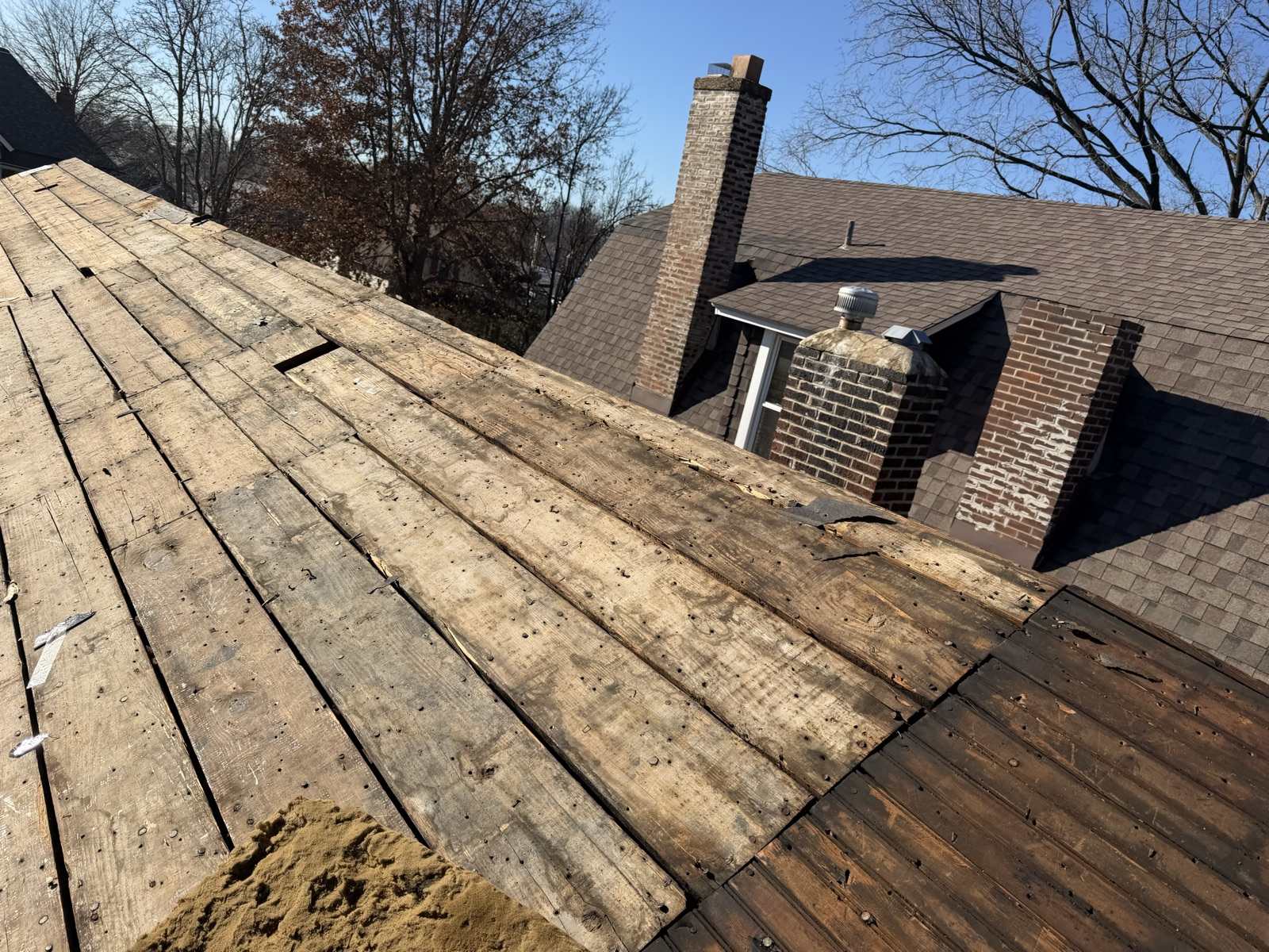 A roof plane on a Keys Roofing job stripped to its original wide plank decking, boards weathered, split at the edges and separated by open gaps, studded with old nail shanks, with a pile of loose granules at the lower left. Beyond it the main house still carries its weathered brown shingles, with two brick chimneys showing spalled brick and patched mortar and a dormer with a metal turbine vent under a clear blue sky.