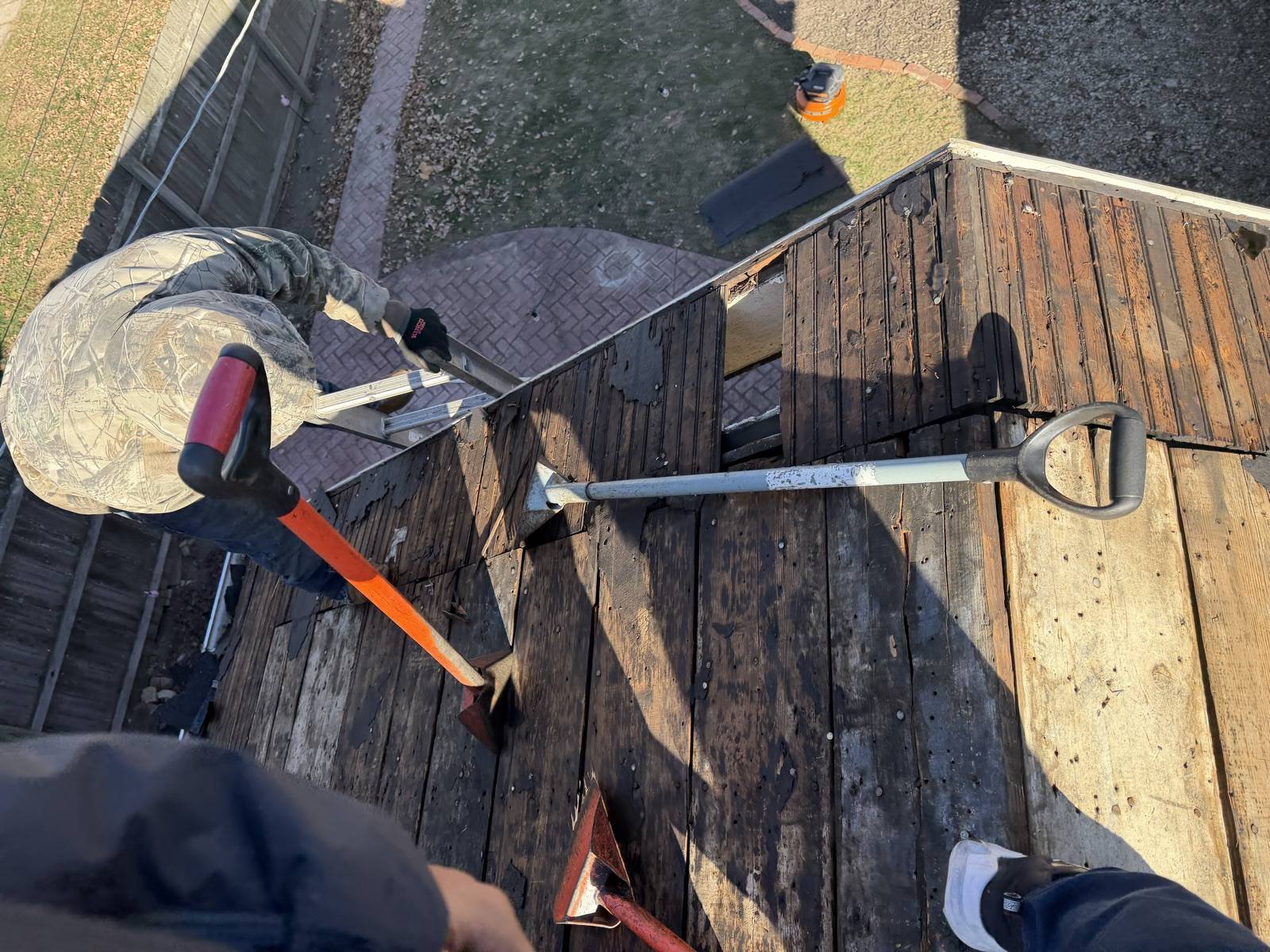 Looking down from a roof at dark stripped plank decking on a Keys Roofing job, with an orange handled tear off bar, an aluminium D handle roofing shovel and two red flat shovels lying across the boards, an aluminium ladder hooked over the eave, a worker in a camouflage jacket and gloves crouched at the roof edge, and a brick paver patio, timber fence and lawn below.